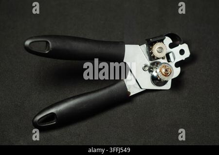 Handy can opener rests on dark surface. Black handles contrast with metallic mechanism. Stock Photo