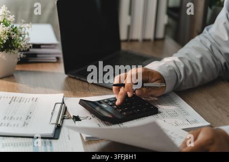 Businessman calculating figures on printed financial documents with calculator, pen, laptop for corporate budgeting, accounting review, revenue planni Stock Photo