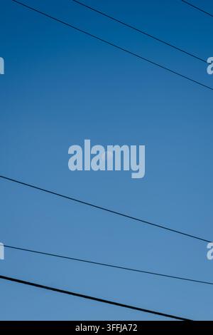 Clear blue sky holds a faint, silvery moon. Power lines stretch across the frame, stark and dark. Moon hangs quietly, almost lost in vastness. Stillne Stock Photo