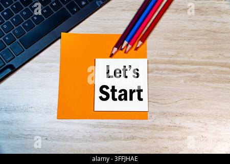 Let's Start text on sticky note placed on laptop keyboard background with colorful pencils on wooden office desk. Begin project concept. Stock Photo