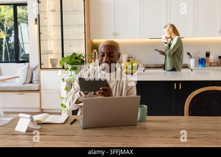 Diverse couple sitting at table in kitchen, man using tablet with laptop, woman holding smartphone Stock Photo