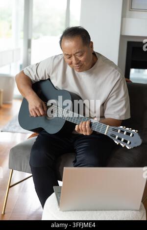 Senior Asian man sitting in home on gray armchair playing black classical guitar following laptop Stock Photo