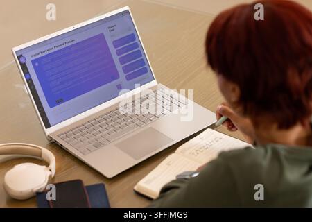 Woman sitting at wooden table dark green top writing in notebook, viewing laptop chat, copy space Stock Photo