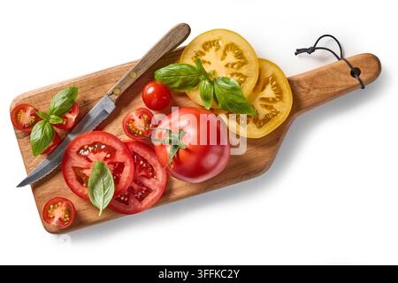 Assortment of fresh colorful whole and sliced tomatoes with green basil leaves arranged on a wooden kitchen cutting board with a knife, photographed f Stock Photo