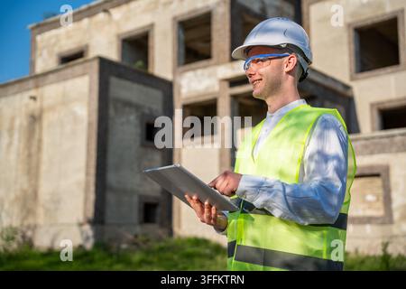 Construction site manager using a laptop beside an unfinished building while wearing safety helmet and high visibility vest. Digital inspection field Stock Photo
