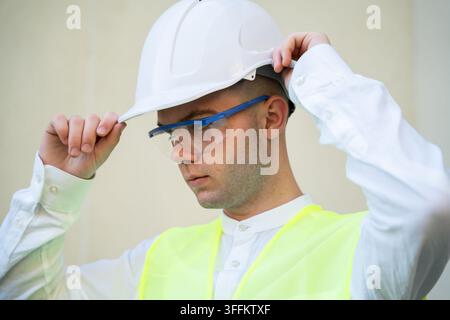 Architect adjusting white hard hat while wearing protective glasses and high visibility vest at building site. Construction safety personal protective Stock Photo