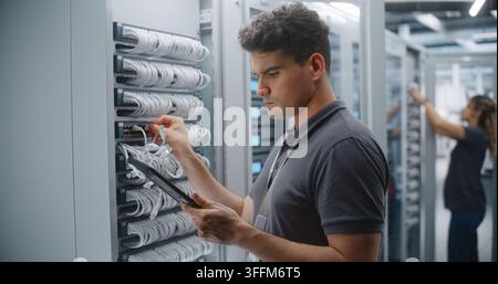 Male IT Engineer Checks Rack Connections While Consulting Tablet, Verifying Physical Cabling Against Network Records During Structured Infrastructure Stock Photo