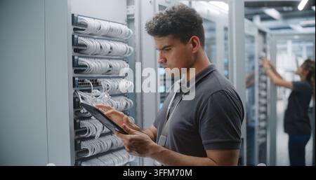 Male IT Engineer Checks Rack Connections While Consulting Tablet, Verifying Physical Cabling Against Network Records During Structured Infrastructure Stock Photo