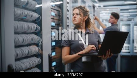 IT Specialist Uses Laptop to Inspect Server Rack Indicators, Male Technician Maintains Cabinet Nearby. Professional Team Performs System Monitoring, I Stock Photo