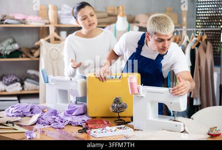 Asian woman dressmaker called a master to repair a sewing machine. Mechanic repairs a sewing machine next to a seamstress in the workshop Stock Photo