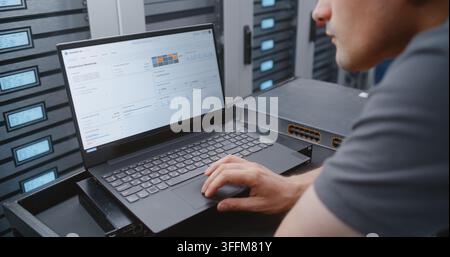 IT Administrator Uses Laptop Beside Network Switch to Navigate Server, Network and Security Logs, Comparing Software Metrics with Hardware Status Thro Stock Photo