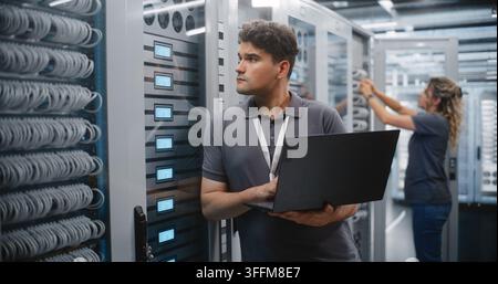 IT Engineer Uses Laptop to Monitor Server Rack Indicators in Data Center, Female Technician Maintains Another Cabinet. Professional Team Performs Cont Stock Photo