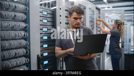 IT Engineer Uses Laptop to Monitor Server Rack Indicators in Data Center, Female Technician Maintains Another Cabinet. Professional Team Performs Cont Stock Photo
