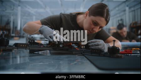 Young Female Specialist Wearing Protective Gloves Uses Precision Screwdriver Assembling FPV Drone Frame. Focused Woman Connects Electronic Flight Cont Stock Photo