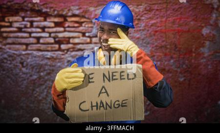 Man in blue hardhat and yellow ear defenders holding a cardboard sign reading 'we need a change' while touching his face with a gloved hand on a stree Stock Photo