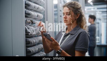 Female IT Specialist Checks Ethernet Connections and Verifies Rack Topology on Smartphone, Matching Physical Ports With Digital Records While Male Technician Works on Another Server Cabinet. Stock Photo