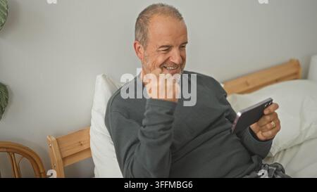 Man holding smartphone with right hand, fist pump gesture while seated propped against pillows on wooden bed frame in a building; joy relaxation conte Stock Photo