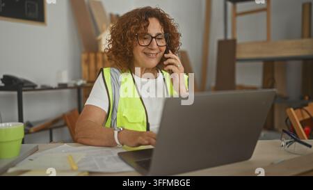 Woman carpenter holds phone to ear and types on laptop amid wood boards and safety glasses in a studio; focused planning. Stock Photo