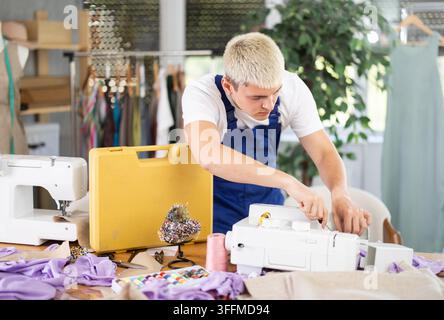 Repairman repairs a sewing machine in the workshop against the background of racks with sewing accessories. Master checks the motor of sewing machine Stock Photo