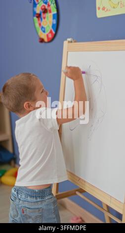 Boy drawing on whiteboard at playroom easel with marker in hand, wearing white t shirt and jeans; concentration creativity. Stock Photo