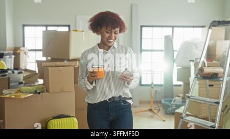 Woman holds smartphone and orange mug, hand tapping phone while surrounded by moving boxes in building; settling in contentment. Stock Photo