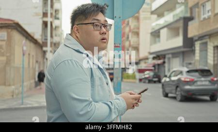 Man in blue shirt holding smartphone and checking the screen while standing by a street sign on a city street with glasses and plus size profile; curi Stock Photo
