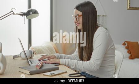 Young woman opens laptop on bedroom desk while smiling and reaching for screen; focused study productivity. Stock Photo