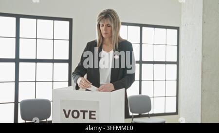 Woman inserting ballot into transparent ballot box with sticker visible in polling building interior, american flags and 'every vote counts' sign; civ Stock Photo