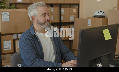 Man typing on computer with packages and stacked boxes, hands on keyboard in a packing area inside a building; focused efficiency. Stock Photo