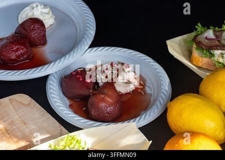 Dark red poached pears served in syrup on a white plastic plate, garnished with cream, sunflower seeds, and crushed freeze-dried berries, positioned n Stock Photo