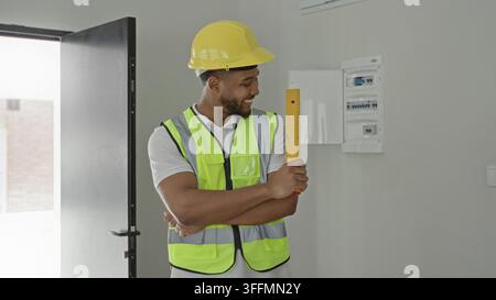 Man in yellow hardhat and hi vis vest holds yellow measuring ruler with crossed arms and smiles inside building; professional confidence. Stock Photo