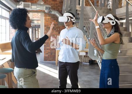 Diverse coworkers testing two white VR headsets in loft-style office with tablet, staircase, ducts Stock Photo