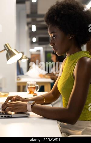 Diverse female coworkers typing on laptop at open-plan office with brass lamp, smartwatch Stock Photo