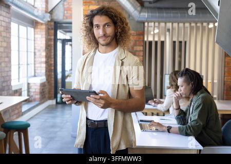 Diverse coworkers working in open-plan office, man holding tablet while colleagues using laptops Stock Photo