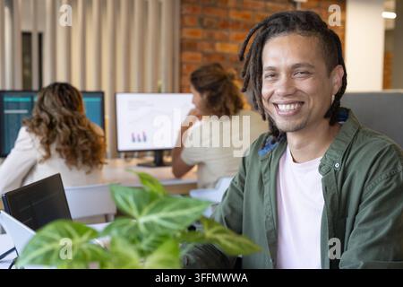 Diverse coworkers working in open-plan office, using monitors, laptop, keyboard, potted plant Stock Photo
