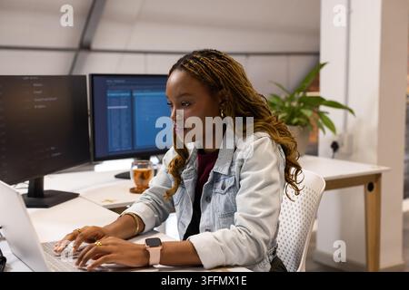 African American woman typing on laptop in denim jacket at office desk with dual monitors, mug Stock Photo