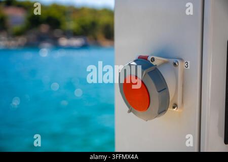 Electrical control switch mounted on a white panel of Electric charging station for boats near a turquoise sea, blurred boats and greenery in the Stock Photo