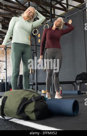 Female friends stretching necks in warehouse gym with blue mat, green duffel, rings Stock Photo