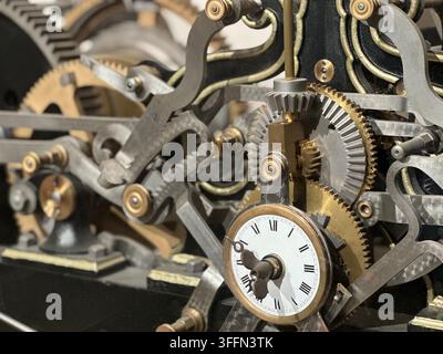 Vintage clockwork mechanism with brass gears, cogs, and a white Roman numeral dial. Intricate brass and steel clockwork gears surround a white clock f Stock Photo