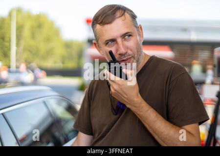 Man Using Handheld Radio Beside a Car. Middle-aged man holding a handheld two-way radio near his mouth while standing beside a car outdoors. Stock Photo