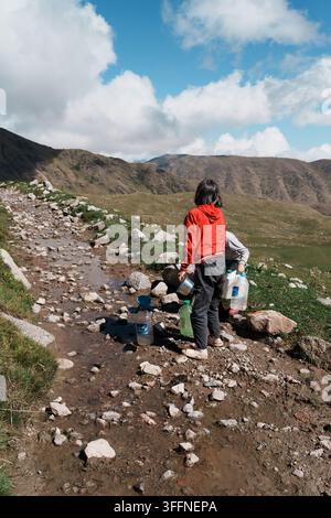 Hiker in red jacket stands on rocky trail, facing distant mountains. Sky blue with fluffy clouds above. Person appears focused, preparing gear or rest Stock Photo