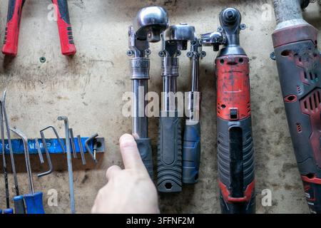 Car mechanic picking up a ratchet from a collection of hand and power tools organised on a workshop wall. The tool display features a mixture of tools Stock Photo