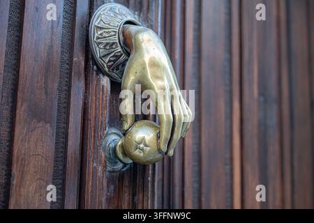 Close-up of an ornate brass door knocker featuring a sculpted hand holding a spherical handle, mounted on a richly textured dark wooden door in Ávila, Stock Photo