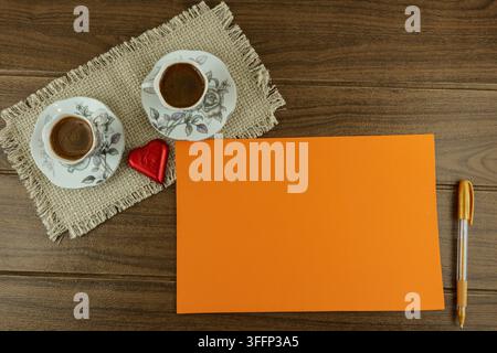 Top view flat lay of a workspace or breakfast table. Two cups of espresso, a blank orange card for text, and red heart-shaped chocolates. Stock Photo