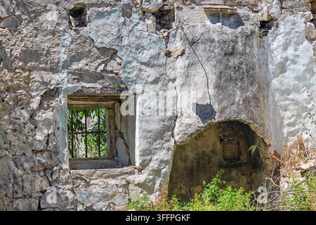 An aged plaster wall of a ruined building featuring a barred window, stone niche and peeling textures, partially reclaimed by overgrown greenery and s Stock Photo
