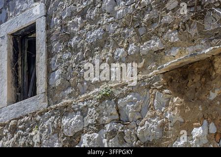 Weathered stone wall with an old wooden window frame and rustic mortar texture, showing cracks, climbing vines and sunlit shadows on an abandoned hist Stock Photo