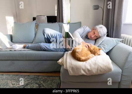Senior man relaxes on a sofa at home, reading a book while gently petting a sleeping ginger cat curled on a soft blanket in a bright, comfortable livi Stock Photo
