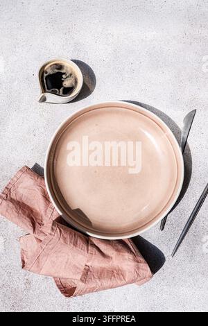 Overhead still life of an empty blush pink ceramic plate beside a dark spoon, used coffee cup and crumpled napkin on a pale textured tabletop with str Stock Photo
