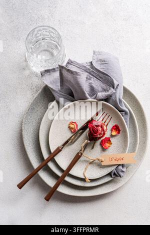 Overhead view of a romantic table place setting with dry red rose petals, ceramic plates, vintage cutlery, gray napkin, glass, and a handmade love tag Stock Photo