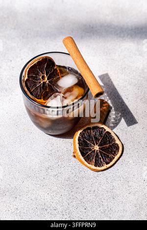 Glass of spiced iced coffee with melting ice, cinnamon stick, and dried orange slices on a bright textured surface, captured in natural sunlight with Stock Photo